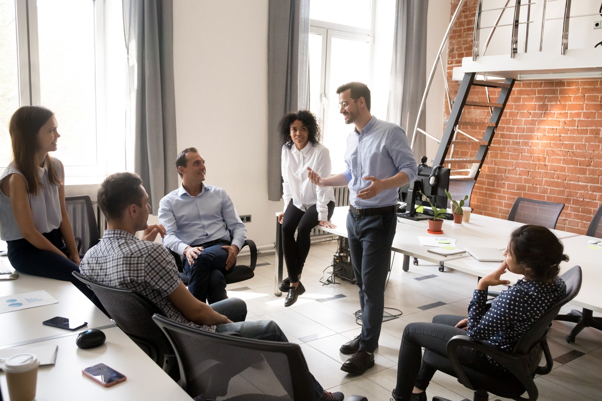 Confident male leader, coach talking with multiracial group of office workers, having good conversation with subordinate, brainstorming, discussing business strategy, ideas, team building activity.